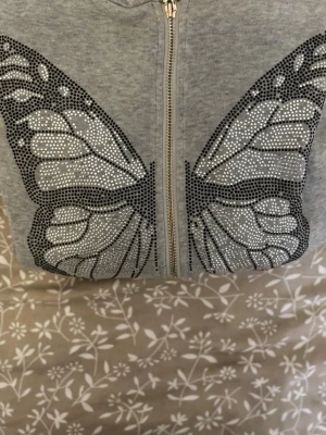 Grå hoodie med fjärilsdetaljer - Snygg grå hoodie med dragkedja och stora fjärilsvingar i svarta och silvriga stenar på framsidan. Hoodien har långa ärmar, fickor framtill och ribbade muddar. Perfekt för dig som gillar unika detaljer och vill sticka ut lite extra.