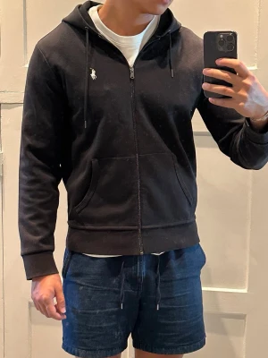 Svart hoodie med dragkedja från Polo Ralph Lauren - Snygg svart hoodie med dragkedja från Polo Ralph Lauren. Klassisk design med huva och dragsnören, två fickor framtill och den ikoniska vita loggan broderad på bröstet. Perfekt till jeans eller shorts för en avslappnad look.