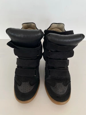 Svarta höga sneakers med kardborreband - Säljer ett par svarta sneakers med högt skaft och flera breda kardborreband. Skorna har en mix av mocka och läderdetaljer samt en brun gummisula. Insidan är ljus och tungan är extra vadderad för en chunky look.