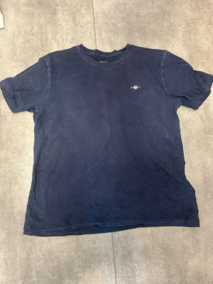 Mörkblå t-shirt från GANT - En enkel och stilren mörkblå t-shirt från GANT med liten logga på bröstet. Klassisk rund halsringning och korta ärmar. Tillverkad i mjuk bomull som känns skön mot huden. Perfekt till jeans eller shorts för en clean look.