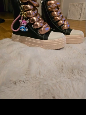 Chunky svarta sneakers med metallic snören - Unika höga sneakers i svart med tjocka metallic-rosa snören och vit tå. Skorna har platåsula i beige och detaljer som en rosa nyckelring med figur. Materialet är en mix av läderimitation och textil. Perfekta för dig som vill sticka ut med din stil.