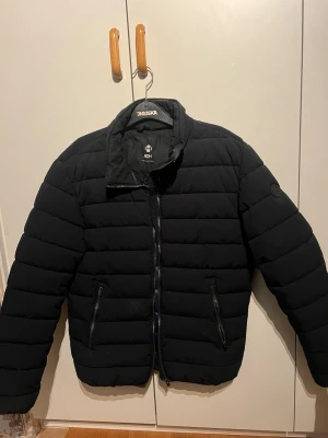 Svart pufferjacka från ADH - Säljer en svart pufferjacka från ADH med hög krage och dragkedja framtill. Jackan har två sidofickor med dragkedja och quiltad design som ger en schysst look. Perfekt för kalla dagar när du vill hålla dig varm och stilren.