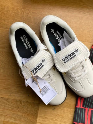 Adidas Wales Bonner vita sneakers - Unika vita Adidas Wales Bonner sneakers med guldfärgade nitar längs sidorna och bred kardborre över snörningen. Skorna har svart sula och detaljer i läder och textil. Perfekt för dig som vill sticka ut med en stilren men ändå vågad sneaker.