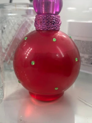 Rund rosa parfymflaska med gröna stenar - En parfym i en rund, klarröd flaska med små gröna stenar och ett lila-glittrigt halsband samt lila kork. Flaskan har en lekfull och färgstark design som sticker ut på sminkbordet. Perfekt för dig som gillar unika och färgglada parfymer.