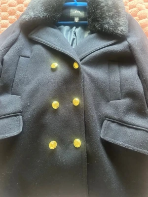 Marinblå skepparkavaj Viva Couture - Stilren marinblå skepparkavaj från Viva Couture med dubbelknäppning och sex guldknappar framtill. Kavajen har bred krage med mjuk fuskpäls, två fickor med lock och är helfodrad. Perfekt för dig som vill ha en klassisk look med en twist.
