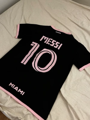 Inter Miami Messi matchtröja svart/rosa - Säljer en svart Inter Miami matchtröja med rosa detaljer, Messi och nummer 10 på ryggen. Framsidan har klubbmärke och sponsorlogga i rosa. Tröjan har korta ärmar och V-ringning, perfekt för fotboll eller streetwear.