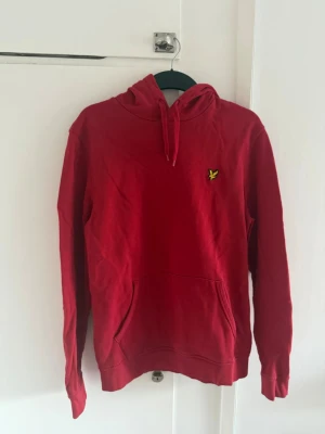 Röd Lyle & Scott hoodie - Snygg röd hoodie med huva och dragsnören. Har en gul broderad logga på bröstet och en stor känguruficka framtill. Tillverkad i mjukt bomullstyg och har ribbade muddar vid ärmar och nederkant. Perfekt för en chill och avslappnad stil.