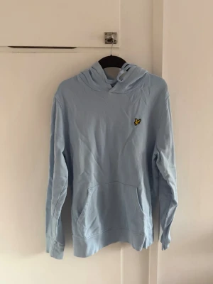 Ljusblå Lyle & Scott hoodie  - Snygg ljusblå hoodie med huva och känguruficka framtill. På bröstet sitter en liten gul logga som detalj. Hoodien har långa ärmar och är gjord i mjukt bomullstyg, perfekt för en avslappnad stil. Har en liten gul fläck på bröstet/magen