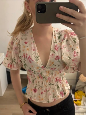 Blommig omlottopp med puffärm - Supersöt blommig omlottopp med puffärmar och smock i midjan. Toppen har v-ringning och är croppad, perfekt till höga jeans eller kjol. Färgerna går i vitt, rosa, grön och gul med ett romantiskt blommönster.