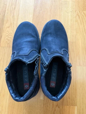 Svarta Chelsea boots från Zeus - Snygga svarta Chelsea boots från Zeus med dragkedja på sidan och kontrastsömmar. Skorna har en klassisk rund tå och är tillverkade i ett slitstarkt material som ser ut som läder. Perfekta för dig som vill ha en stilren och enkel look.