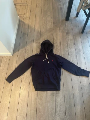 Mörkblå hoodie med huva - Snygg mörkblå hoodie med huva och vita dragsnören. Klassisk känguruficka framtill och en liten röd logga broderad på bröstet. Hoodien har långa ärmar och ribbade muddar vid ärmslut och nederkant. Perfekt för en avslappnad stil. Pris kan diskuteras . Och kom privat för fler frågor