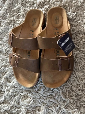 Bruna Birkenstock sandaler - Klassiska bruna sandaler från Birkenstock med två breda remmar och justerbara metallspännen. Tillverkade i läder med mjuk fotbädd för extra komfort. Perfekta för en avslappnad och stilren look, inte äkta men ser bra ut ändå, passar mer storlek 39 än 41.