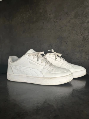 Puma sneakers - Säljer ett par klassiska vita sneakers från Puma i lågt snitt. Skorna har en stilren design med vita snören, diskret Puma-logga på sidan och slitstark gummisula. Tillverkade i syntetmaterial och läderimitation, perfekta för en clean och sportig look.