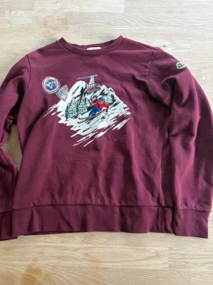 Vinröd moncler hoodie - Vinröd sweatshirt med tryck av skidåkare och berg på framsidan samt broderade märken på bröstet och ärmen. Rund hals och långa ärmar. 
