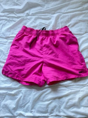 Rosa badbyxor från Nike - Säljer ett par knallrosa shorts från Nike med svart snörning i midjan och liten svart logga på benet. De har en relaxed passform och elastisk midja, med nät i.