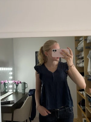 Mörkblå ärmlös blus med volang - Säljer en mörkblå blus med v-ringning, knappar framtill och volangdetaljer vid axlarna. Blusen är ärmlös och har en luftig, lite loose passform. Materialet känns mjukt och har en lätt struktur. Perfekt för dig som gillar en enkel men söt topp.