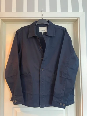 Mörkblå overshirt från !Solid - Stilren mörkblå overshirt från !Solid med klassisk krage, knäppning framtill och två stora fickor. Overshirten är gjord i ett slitstarkt bomullstyg och har knappar vid ärmsluten för en snygg finish. Perfekt lager på lager-plagg för en clean look. Tröjan är helt ny och är aldrig använd. NYPRIS 800kr