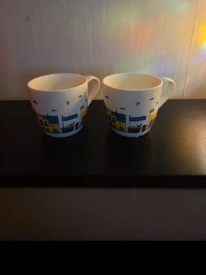 Två muggar med Sverige-motiv - Säljer två vita porslinsmuggar med färgglatt motiv av svenska hus, flaggor och fåglar. På ena sidan står det 'Sweden'. Perfekta för te eller kaffe och har bekvämt handtag. Muggarna har en lekfull och modern design som sticker ut i köket.