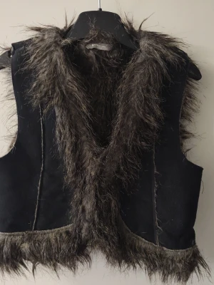 Svart pälsväst tjej stl 134/140 - Cool svart väst med lång, mörkbrun fuskpäls längs krage, framkant och nederkant. Västen är ärmlös och har en trendig, lite ruffig look som passar perfekt till lager-på-lager-stil. Materialet är mjukt och pälsen ger en lyxig känsla. Tjej stl 134/140. Bra skick. Fuskpäls.