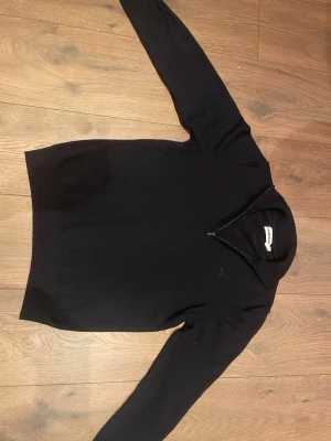 Svart halvzip-tröja från J.Lindeberg - Stilren svart tröja från J.Lindeberg med halv dragkedja och ribbad krage. Tröjan har lång ärm och diskret logga på bröstet. Perfekt för en snygg och enkel look. Materialet känns mjukt och bekvämt.
