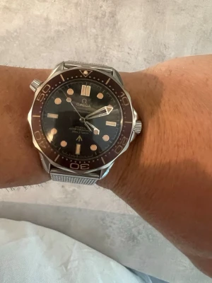 Omega Seamaster klocka i silver - Säljer en Omega Seamaster klocka med mörk urtavla och mesh-armband i silver. Klockan har tydliga indexmarkeringar, vridbar bezel och robust boett i metall. Perfekt för dig som gillar klassisk och sportig stil.
