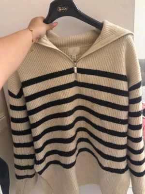 Beige stickad tröja med svarta ränder - Snygg stickad tröja i beige med svarta horisontella ränder. Tröjan har halv dragkedja framtill och en bred krage. Ribbstickad struktur och lång ärm, perfekt för lager-på-lager-stil.