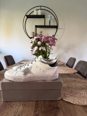 Vita golden goose - Snygga vita sneakers med en silverfärgad stjärna på sidan och grå detaljer. Skorna har vita snören, svart hälparti och en något smutsad sula för en trendig look. Tillverkade i läder med perforerade hål på sidan för ventilation.