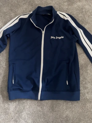Blå zip hoodie från Palm Angels - Snygg marinblå hoodie från Palm Angels med vita ränder längs ärmarna och vit dragkedja. Klassisk logga på bröstet och två fickor med dragkedja. Tillverkad i ett mjukt och stretchigt material som känns sportigt och trendigt. Är ny Scick förutom en liten vitt fläck på högra sidan under bröstet  som knappt syns som ni ser på bilden. Scan och allt sånt funkar 