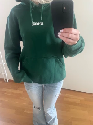 Grön hoodie med tryck - Mörkgrön hoodie med vit text och tryck på både bröstet och ryggen. Huva och stor magficka framtill. Tveka inte att höra av er om frågor eller prisförslag! 