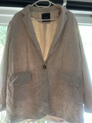 Beige linnékavaj med fickor - Stilren beige kavaj i linne med klassisk krage och en knapp framtill. Två fickor med lock ger en avslappnad men ändå snygg look. Perfekt för dig som gillar en enkel och tidlös stil.
