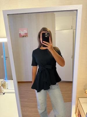 Zara mörkgrönt topp med knyt och kort ärm - Snygg svart topp med korta ärmar och ett brett knytband i midjan som ger en markerad siluett. Toppen har rund halsringning och lätt utsvängd nederdel för en trendig look. Perfekt att styla med jeans eller kjol.