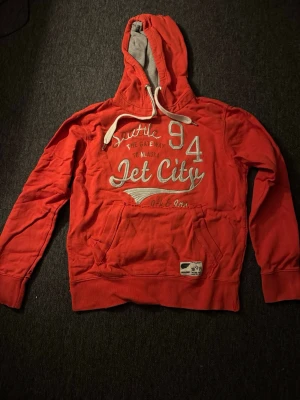 Jack & Jones hoodie - Snygg orange hoodie från Jack & Jones med vit text och tryck på bröstet. Stor känguruficka framtill, vita dragsnören och grå insida i huvan. Tillverkad i mjuk bomullsblandning, perfekt för chill dagar. Stora trycket 'Jet City 94' ger en sportig vibe.