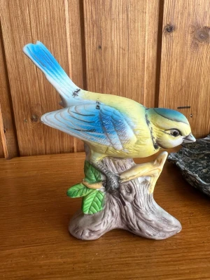 Fågelprydnad i porslin - Detaljerad prydnadsfågel i porslin med blå, gula och vita nyanser. Fågeln sitter på en brun gren med gröna blad och har realistiska former och färgskiftningar. Perfekt som dekoration för dig som gillar natur och djurmotiv.