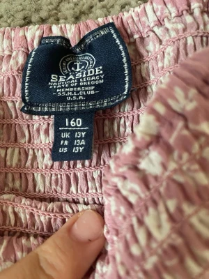 Rosa blommig volangkjol - Söt rosa kortkjol med vita blommor och bred smock i midjan. Kjolen har dubbla volanger som ger en luftig och lekfull känsla. Perfekt för dig som gillar en feminin och avslappnad stil.