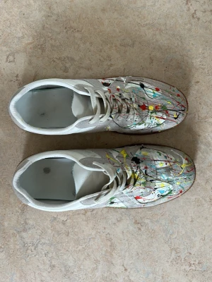 Vita gats med färgstänk - Unika vita sneakers med färgstänk i rött, gult, svart, blått och grönt. Skorna har klassisk låg siluett, vita snören och sula i gummi. Pris kan diskuteras!