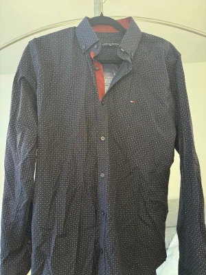 Mörkblå prickig skjorta Tommy Hilfiger - Snygg mörkblå skjorta från Tommy Hilfiger med små vita och röda prickar. Klassisk krage, knappar framtill och diskret logga på bröstet. Perfekt för dig som vill ha en stilren men ändå unik look.
