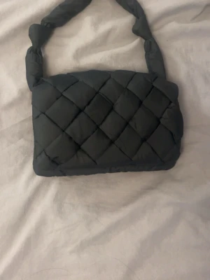 Svart quiltad handväska - Snygg svart quiltad handväska med vadderad axelrem och rutmönster. Väskan har två stora fack och en innerficka med dragkedja. Perfekt för dig som gillar trendiga accessoarer och vill ha plats för allt du behöver.