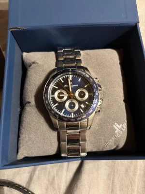 Jacques Lemans silverfärgad klocka - Stilren klocka från Jacques Lemans med silverfärgad länk i metall och mörkblå urtavla. Klockan har tre mindre urtavlor, tydliga markeringar och kronograf-funktion. Levereras i originalask med extra länkar och svart läderarmband.