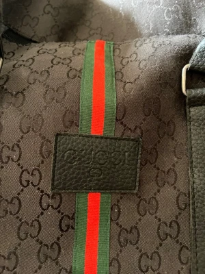 Svart weekendbag från Gucci - Snygg svart weekendbag från Gucci med klassiskt GG-mönster, svarta läderdetaljer och de ikoniska gröna och röda ränderna i mitten. Väskan har två robusta handtag och är perfekt för resor. Materialet är en mix av canvas och läder. För mer bilder kom privat !