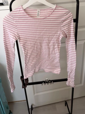 Rosa och vit randig långärmad topp - Säljer en söt långärmad topp med rosa och vita ränder. Toppen har rund halsringning och är gjord i ett mjukt, ribbat bomullsmaterial som sitter tajt mot kroppen. Perfekt för dig som gillar en enkel men fräsch stil.