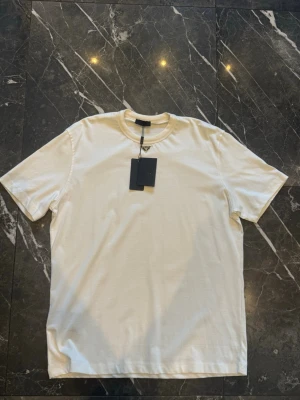 Vit t-shirt från Prada  - Snygg vit t-shirt från Prada med klassisk rund hals och diskret logga vid halsen. Tillverkad i mjuk bomull för en clean och stilren look. Perfekt till jeans eller shorts för en enkel men lyxig vibe.