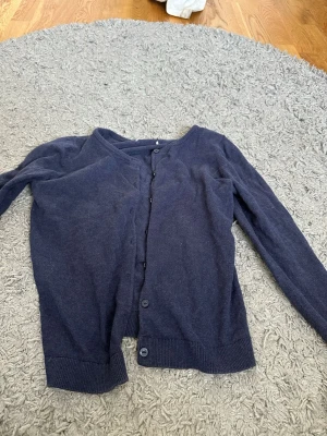 Mörkblå basic kofta från H&M - En enkel och stilren mörkblå kofta med rund halsringning och knäppning framtill från H&M. Koftan har långa ärmar och ribbade muddar vid ärmslut och nederkant. Perfekt att slänga över en topp för en avslappnad look.