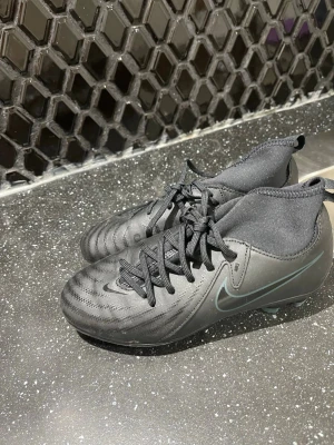 Svarta Nike fotbollsskor med socka - Säljer ett par svarta Nike fotbollsskor med högre strumpliknande skaft och snörning. Skorna har en stilren design med diskret Nike-logga på sidan och är tillverkade i syntetmaterial för bra bollkontroll. Perfekta för dig som vill ha grym touch på planen.