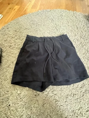 Svarta kostym shorts med snörning från ONLY - Svarta kostym shorts från ONLY med snörning i midjan och bälteshällor. De har en enkel och stilren design, perfekta för varma dagar. Tillverkade i mjukt material som känns skönt mot huden.