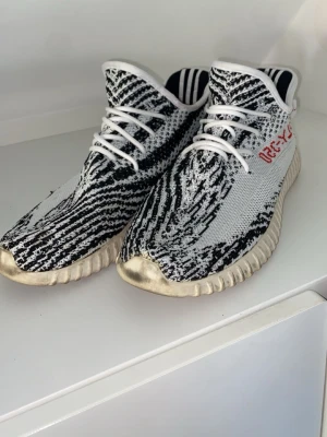 Yeezys 350  - Säljer ett par sneakers med svartvitt zebramönster och vita snören. Skorna har en tjock, räfflad sula i beige och vit samt röda detaljer på sidan. Ovandelen är i stickad textil för en skön och flexibel känsla.