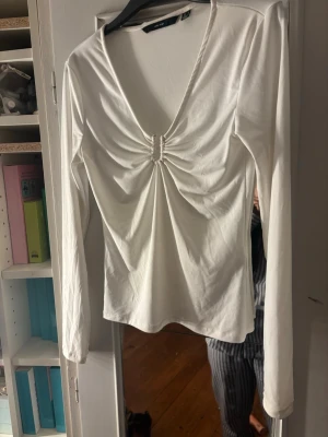 Vit långärmad topp med rynkdetalj - Säljer en vit långärmad topp med djup v-ringning och snygg rynkdetalj framtill. Toppen har en figurnära passform och är tillverkad i mjukt bomullsmaterial. Perfekt att styla med jeans eller kjol för en trendig look.