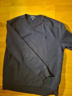 Mörkblå sweatshirt från Polo Ralph Lauren - Klassisk mörkblå sweatshirt från Polo Ralph Lauren med rund halsringning och diskret broderad logga på bröstet. Tröjan har ribbade muddar vid ärmslut och nederkant. Perfekt för en avslappnad och stilren look.