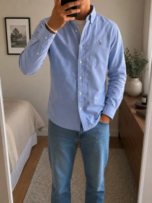 Blå oxfordskjorta från Ralph Lauren - Klassisk ljusblå oxfordskjorta från Ralph Lauren i 100% bomull. Skjortan har button-down krage, långa ärmar och den ikoniska broderade loggan på bröstet. Perfekt till jeans eller chinos för en clean och stilren look.