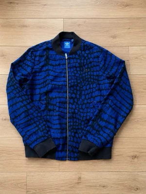 Blåmönstrad bomberjacka från Adidas - Snygg blå bomberjacka från Adidas med svart krokodil-liknande mönster och stor ADIDAS-text på ryggen. Jackan har svarta muddar, dragkedja framtill och klassisk bomberkrage. Perfekt för dig som vill sticka ut med en unik streetwear-look. Storlek S