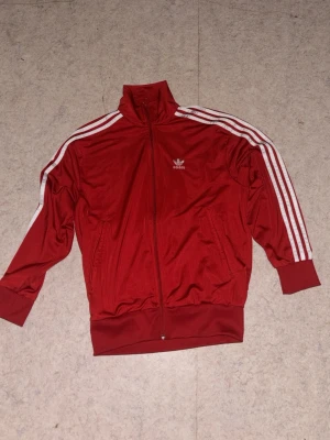 Röd Adidas track jacket - Snygg röd track jacket från Adidas med klassiska vita ränder längs ärmarna och Adidas-logga på bröstet. Dragkedja framtill, hög krage och muddar vid ärmslut och midja. Perfekt för sport eller streetwear. Tillverkad i ett glansigt syntetmaterial.